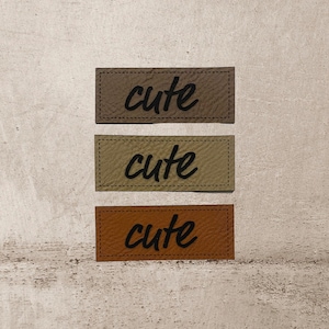 Könnte beinhalten: Drei Leder-Patches mit dem Wort "cute" in schwarzer, kursiv geschriebener Schrift. Die Patches sind in verschiedenen Brauntönen.