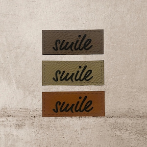 Könnte beinhalten: Drei Leder-Patches mit dem Wort "smile" in schwarzer, kursiv geschriebener Schrift. Die Patches sind braun, grün und orange.