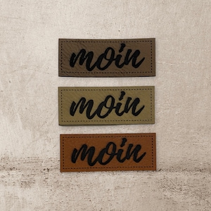 Könnte beinhalten: Drei Leder-Patches mit dem Wort "moin" in schwarzer, kursiv geschriebener Schrift. Die Patches sind in verschiedenen Brauntönen.