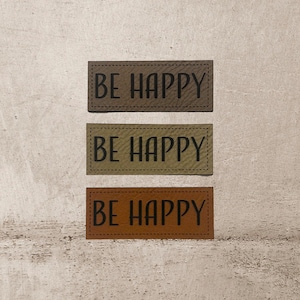 Könnte beinhalten: Drei braune Leder-Patches mit dem Text "BE HAPPY" in schwarzen Buchstaben. Die Patches haben einen genähten Rand.
