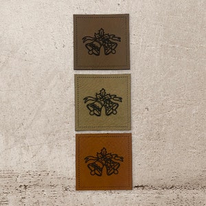 Könnte beinhalten: Drei quadratische Leder-Patches mit einem genähten Rand. Jeder Patch zeigt eine schwarze Umrandung von zwei Glocken mit einem Band und einem Stechpalmenzweig. Die Patches sind braun, beige und orange.
