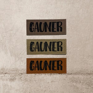Könnte beinhalten: Drei Leder-Patches mit dem Wort "GAUNER" in schwarzer Schrift. Die Patches sind braun, beige und orange.