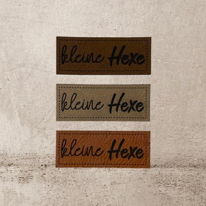 Könnte beinhalten: Drei braune Leder-Etiketten mit dem Text "kleine Hexe" in schwarzer Kursivschrift.