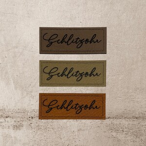 Könnte beinhalten: Drei Leder-Etiketten mit dem Text "Gehlitzohr" in schwarzer Schrift. Die Etiketten sind braun, olivgrün und beige.