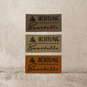 Könnte beinhalten: Drei braune Leder-Etiketten mit schwarzen Nähten und dem Text "ACHTUNG Baustelle" in schwarzen Buchstaben. Die Etiketten sind in verschiedenen Brauntönen.