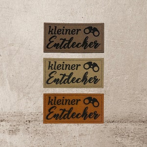 Könnte beinhalten: Drei braune Lederpatches mit dem Text "kleiner Entdecker" und einer schwarzen Silhouette eines Fernglases.