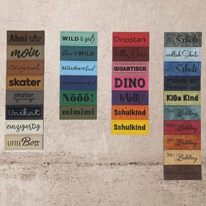 Könnte beinhalten: Eine Sammlung rechteckiger Etiketten in verschiedenen Farben, jeweils mit Text. Die Etiketten zeigen Sätze wie "Schule", "endlich Schule", "Schule", "Vorschulkind", "KiGa Kind", und "Papa's Liebling". Die Etiketten sind für verschiedene Zwecke.