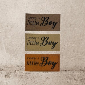 Könnte beinhalten: Drei Stoff-Patches mit einer genähten Kante. Jeder Patch hat eine andere Farbe: Braun, Beige und Orange. Die Patches haben den Text "Daddy's little Boy" in schwarzer Kursivschrift.