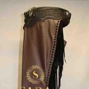Puede incluir: Pantalones de cuero marrón con flecos negros y costuras decorativas. Los pantalones presentan el texto dorado "SARA" y un emblema dorado. Los pantalones están diseñados para montar a caballo y tienen una parte superior decorativa.
