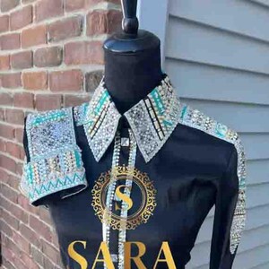 Pode incluir: Camisa preta de manga comprida com gola, punhos e detalhes nos ombros adornados. A camisa apresenta uma frente abotoada e o texto dourado "SARA". A camisa &eacute; exibida em um manequim.