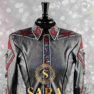 May include: A grey button-up shirt with red and silver decorative accents. The shirt features a pointed collar, long sleeves, and intricate embellishments on the shoulders, cuffs, and placket. The word "SARA" is displayed in gold lettering.