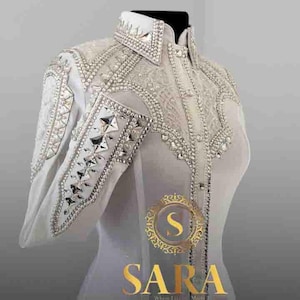 May include: White long-sleeve shirt with intricate silver and crystal embellishments. The shirt features a high collar and detailed patterns on the shoulders and sleeves. The brand name "SARA" is displayed in gold lettering.