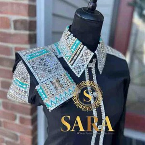 Pode incluir: Camisa preta com gola e detalhes nos ombros adornados. A gola e os ombros apresentam detalhes prateados, turquesas e cristais transparentes. A camisa tem a palavra "SARA" em letras douradas.