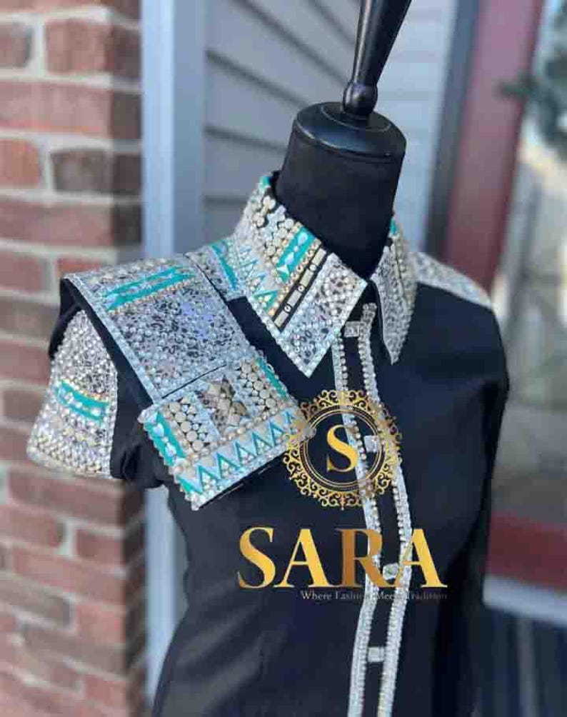 Pode incluir: Camisa preta com gola e detalhes nos ombros adornados. A gola e os ombros apresentam detalhes prateados, turquesas e cristais transparentes. A camisa tem a palavra "SARA" em letras douradas.