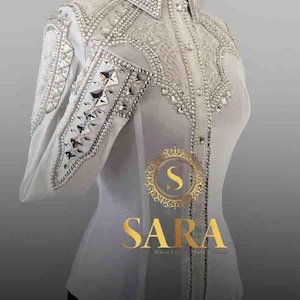 May include: A white, long-sleeved shirt with intricate silver and crystal embellishments. The shirt features a high collar and decorative patterns on the shoulders and sleeves. The brand name "SARA" is displayed in gold lettering.