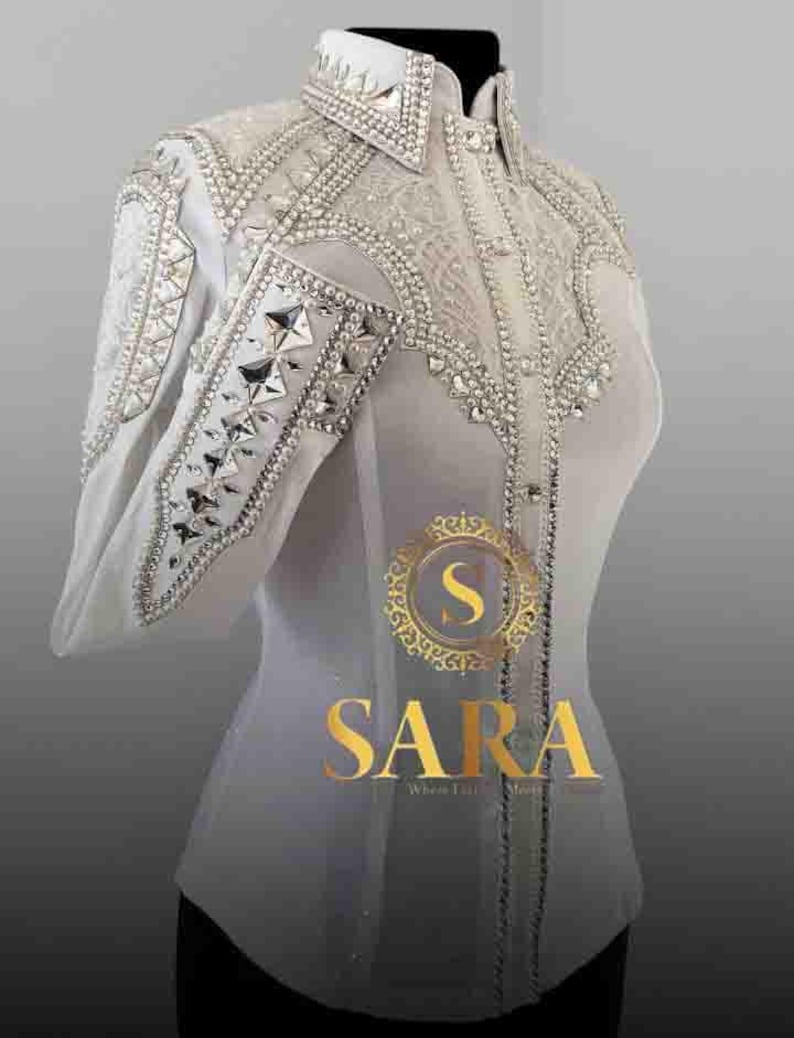 May include: A white, long-sleeved shirt with intricate silver and crystal embellishments. The shirt features a high collar and decorative patterns on the shoulders and sleeves. The brand name "SARA" is displayed in gold lettering.