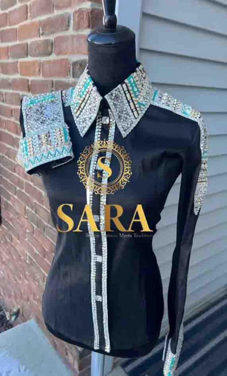 Pode incluir: Camisa preta de manga comprida com gola, punhos e detalhes nos ombros adornados. A camisa apresenta uma frente abotoada e o texto dourado "SARA". A camisa &eacute; exibida em um manequim.