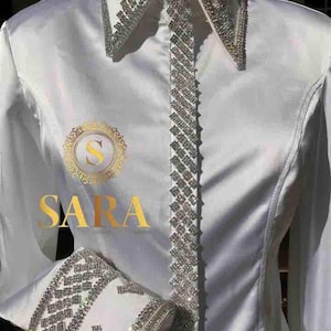 Op de afbeelding: Wit overhemd met lange mouwen met een strassversiering rond de kraag, manchetten en aan de voorkant. Het shirt heeft het woord "SARA" in gouden letters op de voorkant.