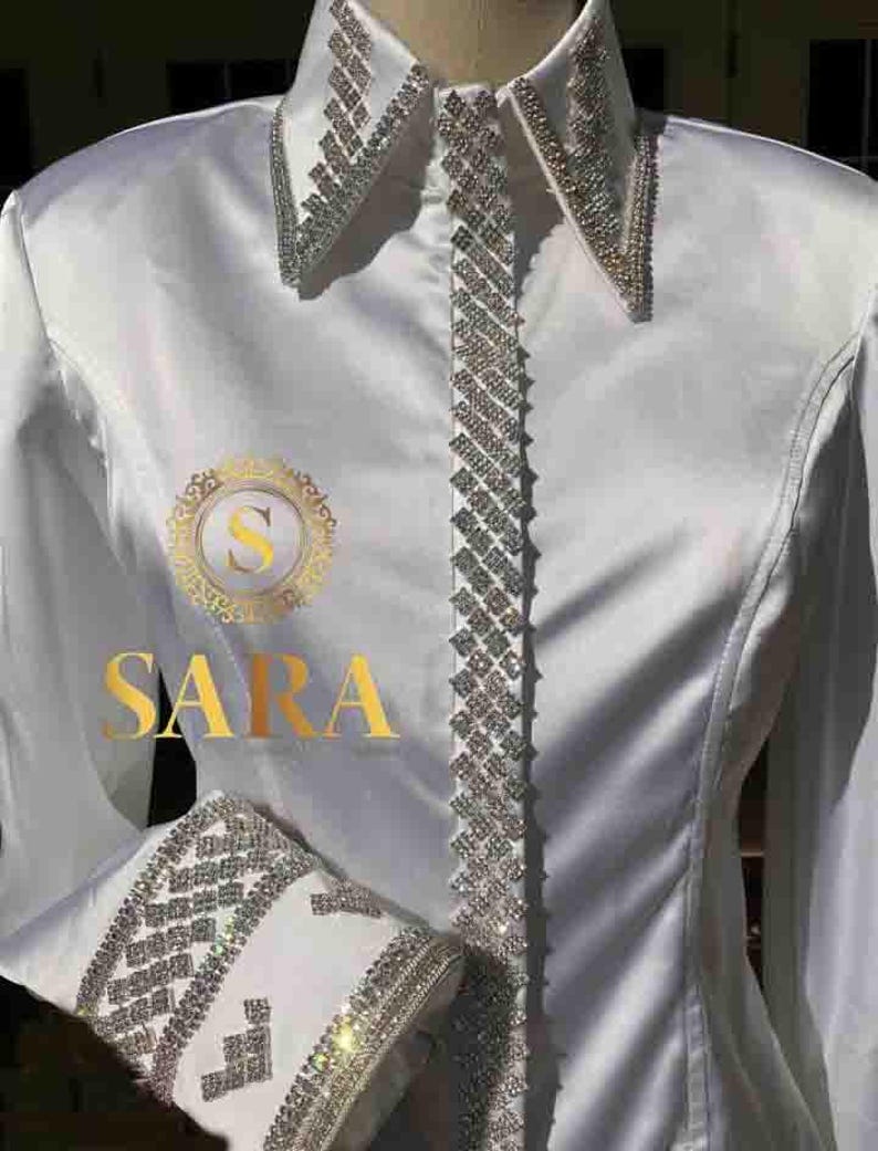 Op de afbeelding: Wit overhemd met lange mouwen met een strassversiering rond de kraag, manchetten en aan de voorkant. Het shirt heeft het woord "SARA" in gouden letters op de voorkant.