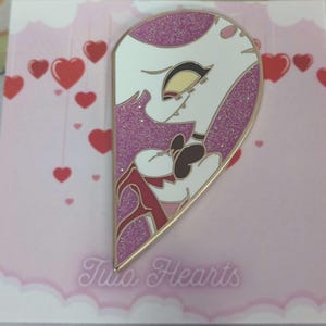 Può includere: Uno spillo in smalto a forma di cuore rosa e bianco con glitter che raffigura il viso di un personaggio dei cartoni animati. Lo spillo dice "Two Hearts" in basso.