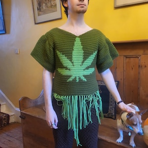 Peut inclure: Un haut crocheté vert avec un grand motif de feuille de marijuana verte et une bordure en franges. Le haut est porté sur un pantalon crocheté marron.
