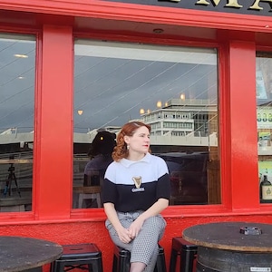 Könnte beinhalten: Eine Frau in einem schwarz-weißen Pullover mit einem Guinness-Logo sitzt auf einem Hocker vor einem roten Gebäude mit einem Schild, auf dem "ERRYMA" steht.