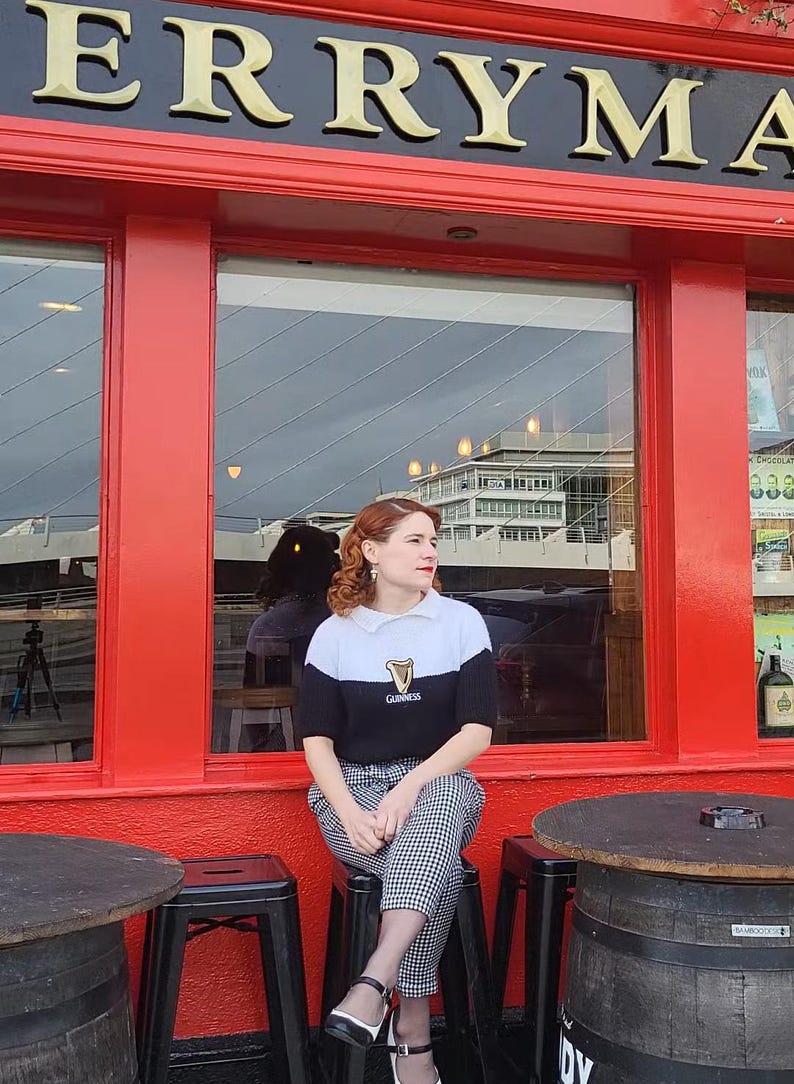 Puede incluir: Una mujer con un su&eacute;ter blanco y negro con el logotipo de Guinness se sienta en un taburete frente a un edificio rojo con un letrero que dice "ERRYMA".