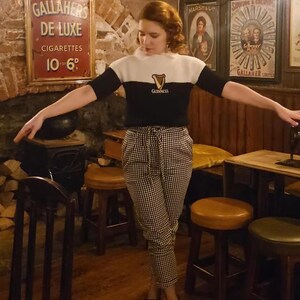 Puede incluir: Una mujer con un su&eacute;ter blanco y negro con un logotipo de Guinness, pantalones a cuadros blanco y negro y zapatos blanco y negro. Est&aacute; de pie en un pub con mesas y sillas de madera.