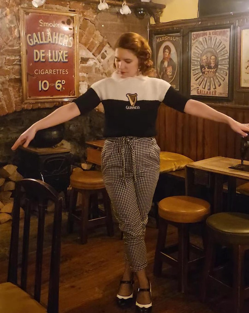 Puede incluir: Una mujer con un su&eacute;ter blanco y negro con un logotipo de Guinness, pantalones a cuadros blanco y negro y zapatos blanco y negro. Est&aacute; de pie en un pub con mesas y sillas de madera.