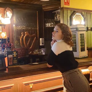 Puede incluir: Una mujer con el pelo rojo que lleva un jersey negro y una camisa blanca con cuello est&aacute; de pie en una barra de bar. Est&aacute; mirando hacia la izquierda y tiene la mano en la barbilla. La barra de bar est&aacute; hecha de madera oscura y tiene un tirador de lat&oacute;n. Hay un vaso de cerveza en la barra.