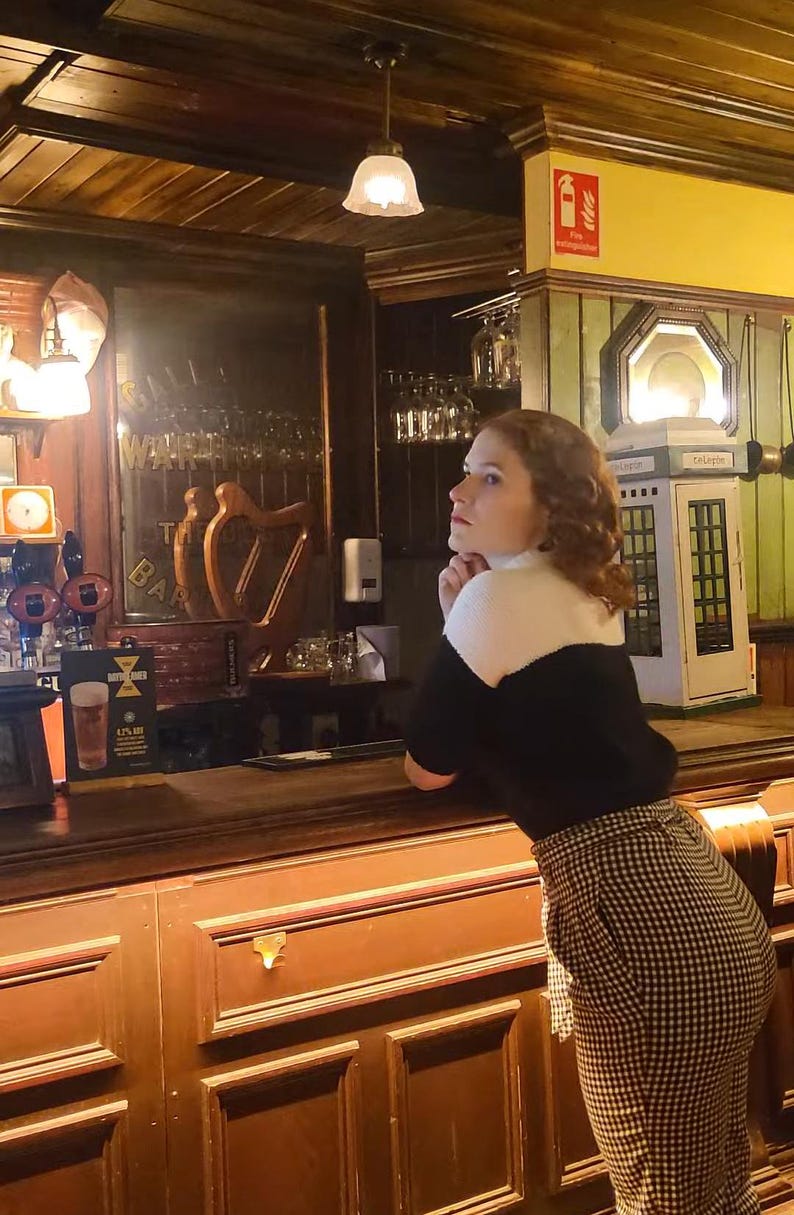 Puede incluir: Una mujer con el pelo rojo que lleva un jersey negro y una camisa blanca con cuello est&aacute; de pie en una barra de bar. Est&aacute; mirando hacia la izquierda y tiene la mano en la barbilla. La barra de bar est&aacute; hecha de madera oscura y tiene un tirador de lat&oacute;n. Hay un vaso de cerveza en la barra.