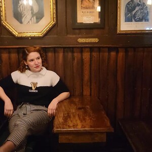 Puede incluir: Una mujer con un su&eacute;ter blanco y negro con la palabra "Guinness" est&aacute; sentada en una mesa de madera en un pub. Las paredes son de madera con obras de arte enmarcadas. La mesa tiene un taburete de cuero marr&oacute;n al lado.