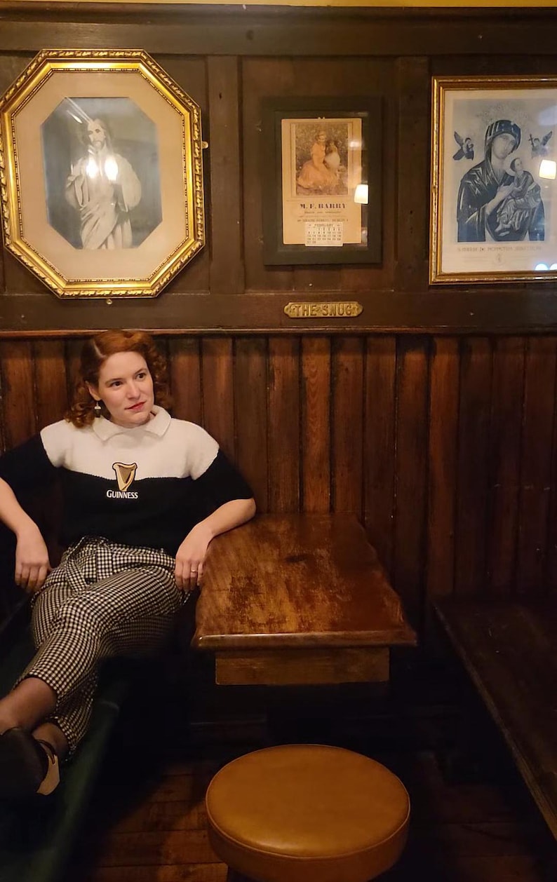 Puede incluir: Una mujer con un su&eacute;ter blanco y negro con la palabra "Guinness" est&aacute; sentada en una mesa de madera en un pub. Las paredes son de madera con obras de arte enmarcadas. La mesa tiene un taburete de cuero marr&oacute;n al lado.