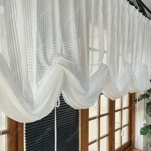 May include: White sheer curtains with a textured, vertical stripe pattern. The curtains are draped over a wooden window frame, creating a soft, flowing aesthetic. The design allows natural light to filter through, enhancing the room's brightness.