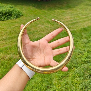 May include: A gold-toned metal choker necklace with rounded edges. The necklace is being held by a hand.