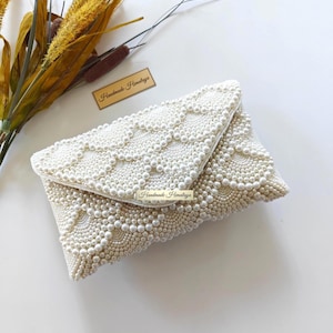 May include: Ivory-colored beaded clutch with a flap closure. The clutch is covered in small, round beads arranged in a decorative pattern. The text "Handmade Himalaya" is visible on a small tag attached to the clutch.