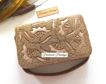 Könnte beinhalten: Eine rechteckige Clutch mit aufwendigen goldenen Perlenstickereien in einem Blumenmuster. Die Oberfläche ist mit metallisch goldenen Perlen bedeckt, die stilisierte Blumenformen bilden. Ein kleines rechteckiges Etikett mit dem Text Handmade Himalaya ist zu sehen.