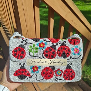 Close-up front view of a handmade ladybug shoulder purse held in hand against wooden stairs. Off-white glass bead background featuring detailed red ladybugs, black spots, green stems, and blue flowers across flap and lower body. White cotton strap visible at sides. Compact seed-bead clutch design with silver hardware snap, offering tactile seed bead texture.