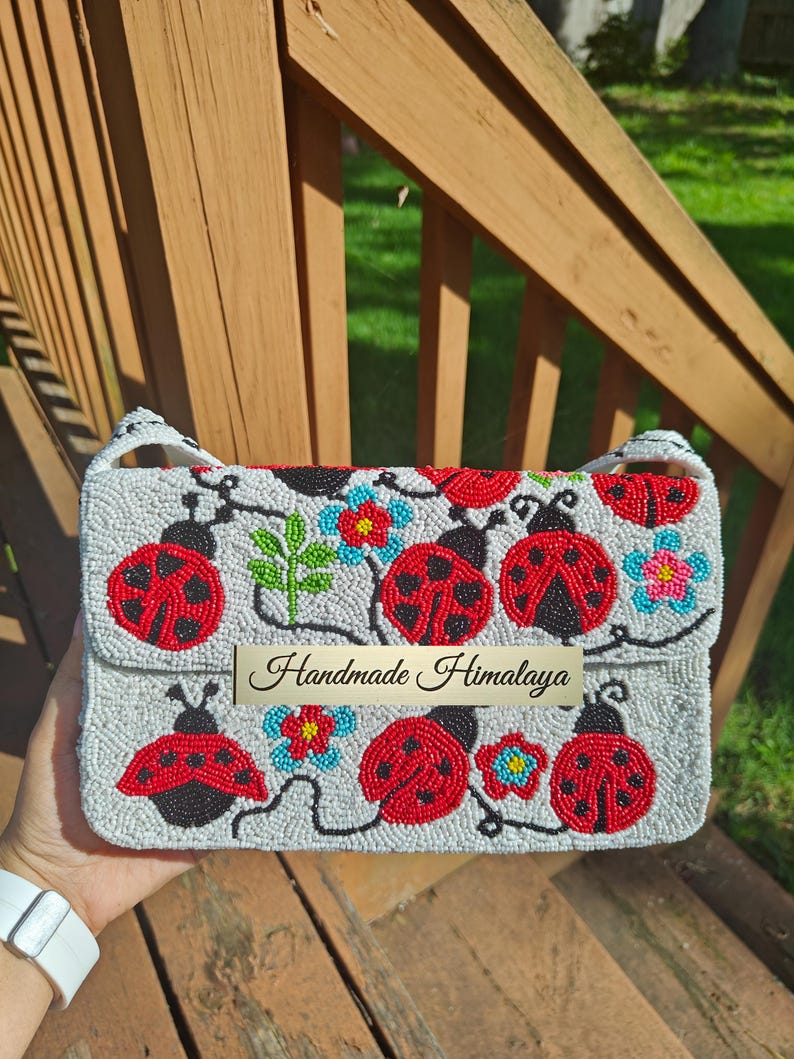 Close-up front view of a handmade ladybug shoulder purse held in hand against wooden stairs. Off-white glass bead background featuring detailed red ladybugs, black spots, green stems, and blue flowers across flap and lower body. White cotton strap visible at sides. Compact seed-bead clutch design with silver hardware snap, offering tactile seed bead texture.