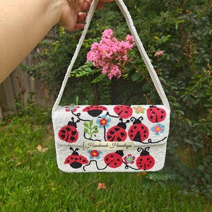 Full front view of a red ladybug beaded bag held up outdoors over a green lawn. Hand-beaded rectangular purse in off-white seed beads featuring red and black ladybugs, green leaves, and colorful flowers. Includes a white beaded shoulder strap attached at top corners. Handmade ladybug clutch style bag with textured seed bead surface and silver snap closure.
