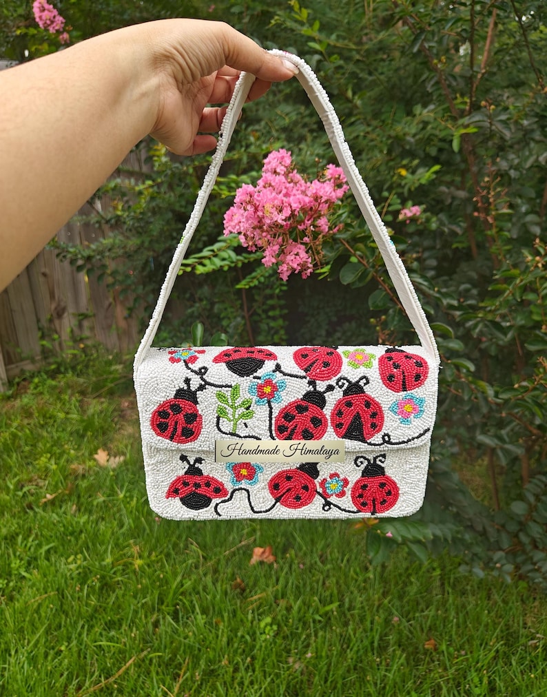 Full front view of a red ladybug beaded bag held up outdoors over a green lawn. Hand-beaded rectangular purse in off-white seed beads featuring red and black ladybugs, green leaves, and colorful flowers. Includes a white beaded shoulder strap attached at top corners. Handmade ladybug clutch style bag with textured seed bead surface and silver snap closure.
