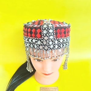 Peut inclure: Un couvre-chef décoratif avec un motif géométrique noir, rouge et blanc. Il est orné d'éléments circulaires argentés et de chaînes pendantes. Le fond est jaune vif.