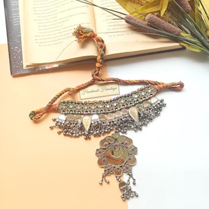 May include: A silver-toned necklace with a large pendant and dangling charms. The necklace has a braided cord with the text "Handmade Himalaya". The pendant has a mirrored center and is surrounded by a floral design with red accents.