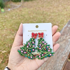 Könnte beinhalten: Ein Paar Weihnachtsbaum-Ohrringe mit grünen Perlen, rosa und goldenen Akzenten und roten Schleifen. Die Ohrringe werden auf einer weißen Karte mit dem Text "HANDMADEHIMALAYA" präsentiert.