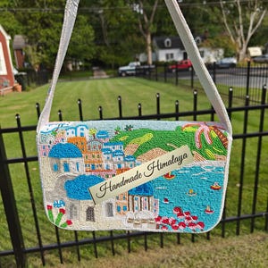 This back view of the handcrafted Santorini beaded bag displays a colorful continuation of the coastal landscape with white and blue Grecian buildings nestled into a green hillside overlooking the bright blue Mediterranean sea. This unique beaded purse captures the spirit of a summer vacation with its artistic beach-inspired beadwork. A stylish accessory for resort fashion and travel, this blue and white handbag makes a special Christmas gift.