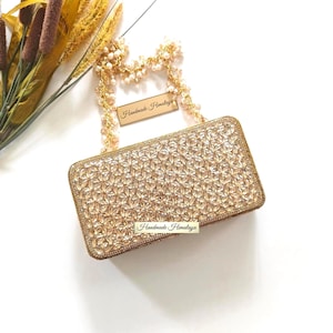 May include: A rectangular gold clutch bag covered in clear, faceted crystals arranged in a floral pattern. The bag features a gold frame and a handle made of gold-toned beads and pearls. A small rectangular tag reads Handmade Himalaya.