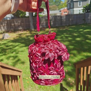 May include: A burgundy-colored, handmade bag with a floral design. The bag features a handle and is adorned with numerous fabric flowers and red beads. The text "Handmade Himalaya" is visible on a small label.