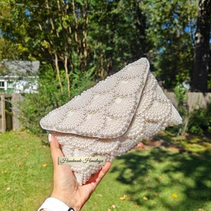 Front view of an ivory pearl beaded wedding clutch held by a hand to demonstrate scale against a lush, sun-drenched garden backdrop. The hand-applied pearls are arranged in a majestic scalloped pattern, transitioning from large center stones to delicate seed beads at the edges. The ivory tones glow with a soft luster, emphasizing the high-density beadwork and the premium "Handmade Himalaya" artisan label.