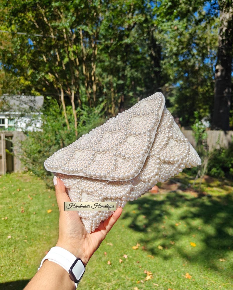 Front view of an ivory pearl beaded wedding clutch held by a hand to demonstrate scale against a lush, sun-drenched garden backdrop. The hand-applied pearls are arranged in a majestic scalloped pattern, transitioning from large center stones to delicate seed beads at the edges. The ivory tones glow with a soft luster, emphasizing the high-density beadwork and the premium "Handmade Himalaya" artisan label.