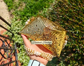 Puede incluir: Un bolso de mano de metal dorado con un intrincado patrón de hojas en relieve y un cierre de bisagra. El bolso cuenta con un asa decorativa hecha de múltiples hilos de pequeñas cuentas blancas y una perla central. Una etiqueta dice Handmade Himalaya.